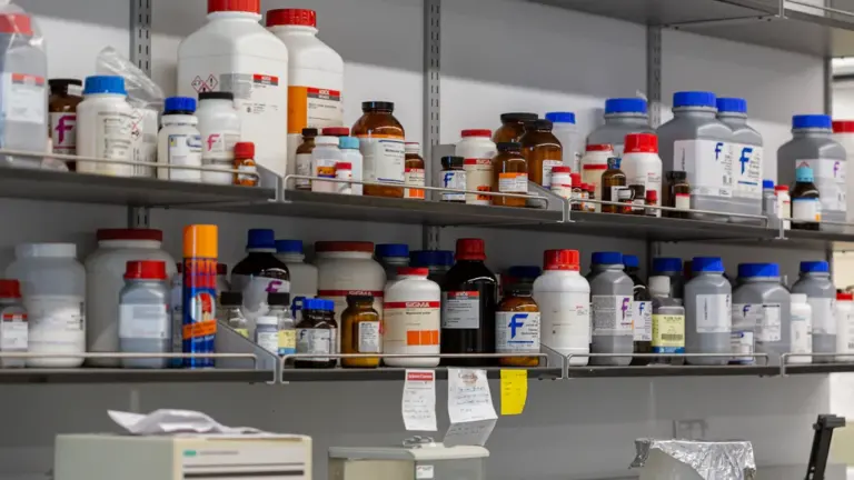 Laboratorijos lentynos su įvairiais cheminiais buteliukais ir aiškiai matomomis etiketėmis.