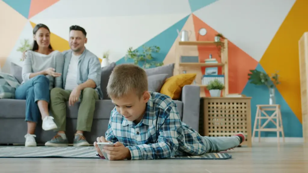 Berniukas žaidžia telefonu svetainėje, o tėvai stebi jį nuo sofos