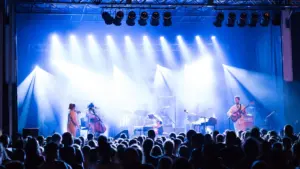 Grupė atlieka muzikos kūrinį scenoje su ryškiu apšvietimu pilnos žiūrovų salės akivaizdoje.