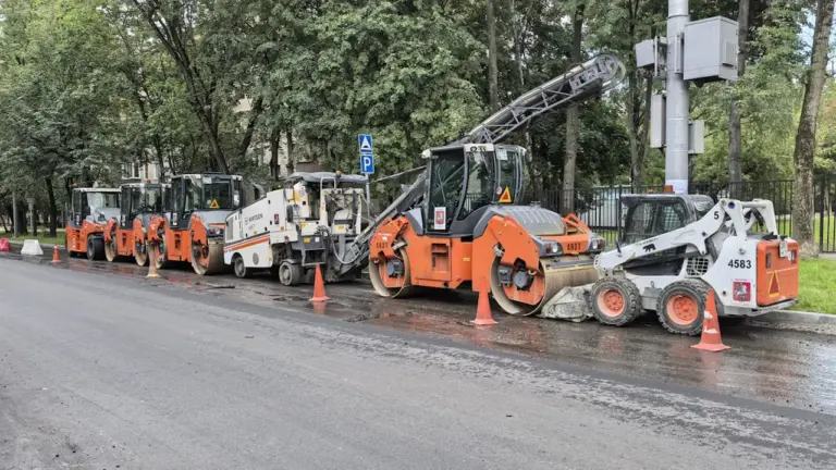 Statybinė technika išrikiuota ant šlapios asfaltuotos kelio dangos remonto darbų metu.