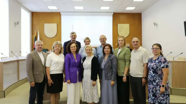 Grupė oficialiai apsirengusių žmonių pozuoja bendrai nuotraukai posėdžių salėje su projektoriumi fone.