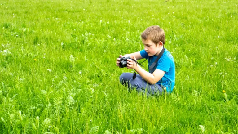 Jaunas berniukas fotografuoja gamtoje su fotoaparatu žaliuojančioje vasaros pievoje.