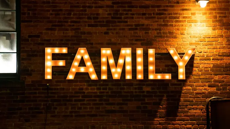Šviečiantis užrašas „FAMILY“ ant raudonų plytų sienos vakare, jaukus kino atmosferos akcentas.