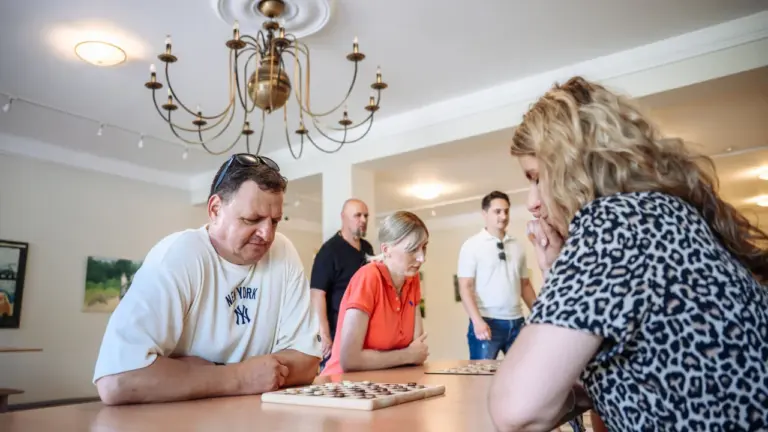 Žmonių grupė susirinkusi prie stalo žaisti šaškėmis šviesioje patalpoje su kabančiu šviestuvu.