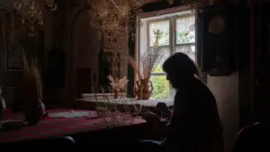 Moteris tamsiame kambaryje prie stalo riša šiaudinius sodus, aplink gausu tradicinių lietuviškų dirbinių.