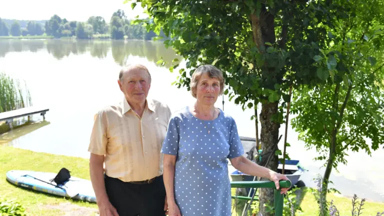 Vyresnio amžiaus pora stovi lauke šalia ežero su medžiais ir valtimis fone.