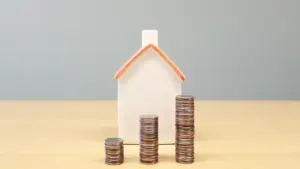 Mažas namo modelis ir monetų stulpeliai ant stalo, rodantys finansinę būsto paskolos situaciją.