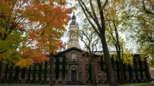 Istorinis universiteto pastatas su laikrodžio bokštu rudeniniame parke, primenantis akademinę aplinką.