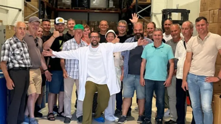 Didelė vyrų ir moterų grupė šypsosi sandėlyje, o vienas vyras viduryje vilki baltą chalatą.