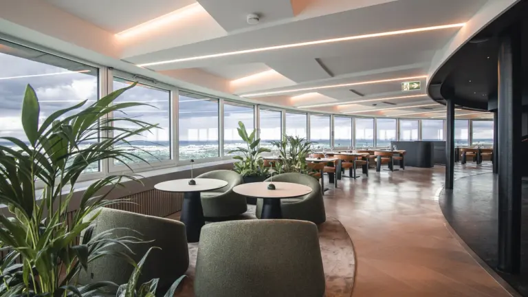 Moderniame restorane su panoraminiais langais stovi jaukūs žali foteliai bei apvalūs staliukai su augalais.