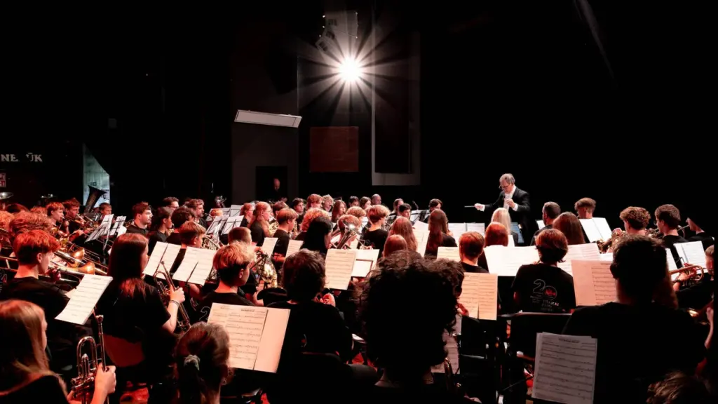 Didelės grupės muzikantų orkestras repetuoja scenoje, vadovaujamas dirigento ryškioje prožektorių šviesoje.