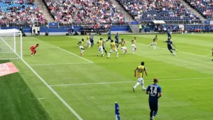 Futbolo rungtynių akimirka stadione, kur žaidėjai varžosi dėl kamuolio kontrolės aikštėje.