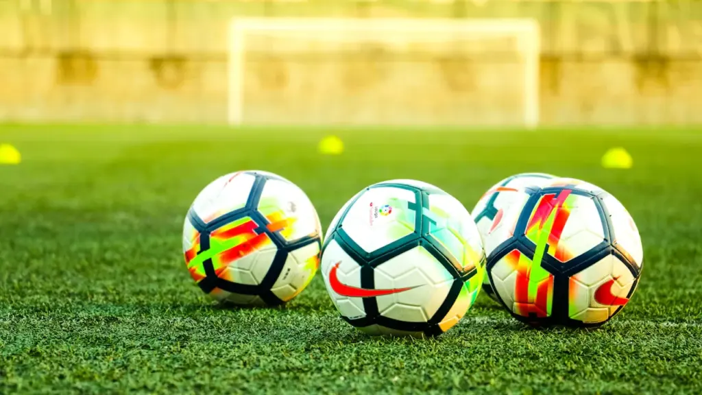 Tryys futbolo kamuoliai išrikiuoti ant futbolo aikštės žolės prieš treniruotę.