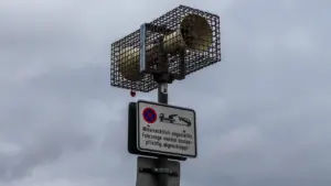 Gatvės garsiakalbis apsauginiame narve pritvirtintas prie stulpo su įspėjamaisiais kelio ženklais.