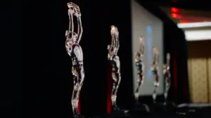 Keli skaidrūs apdovanojimų statulėlės išrikiuoti ant stalo pagerbimo ceremonijos metu.
