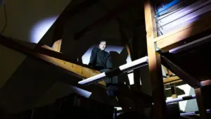 Jaunas vyras tamsiais drabužiais sėdi ant medinės konstrukcijos aukštai po pastato stogu.